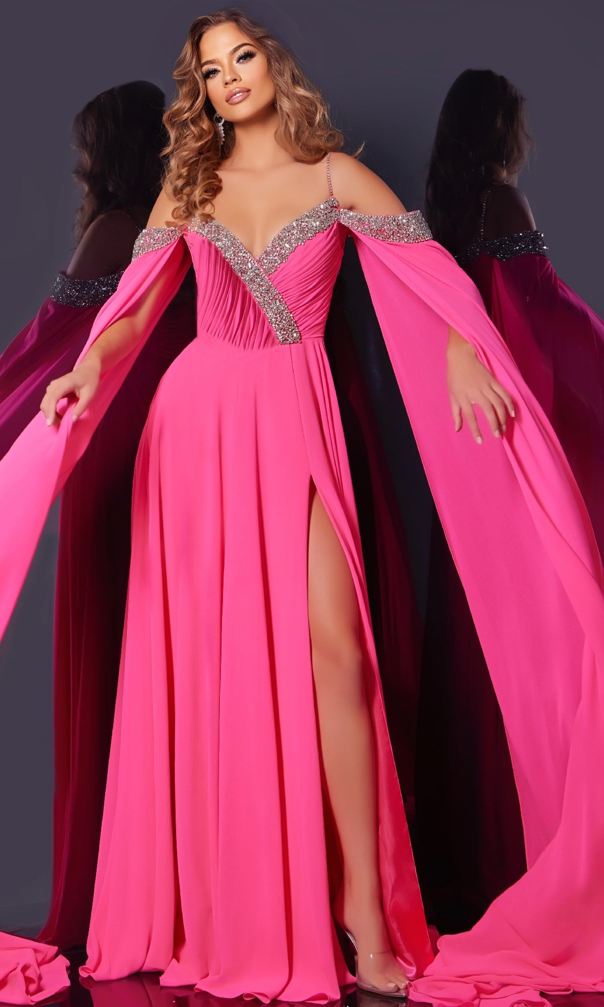 A woman stands against a dark background wearing the Jovani 38996 Long Formal Dress in bright pink, featuring off-shoulder rhinestone details, flowing sleeves, and a thigh-high slit.